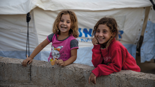 Två leende flickor står framför ett UNHCR-tält.