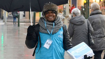 En kille står med ett paraply och UNHCR-väst på ett torg med människor.