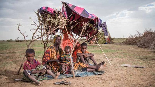 En familj på flykt sitter under en filt som skyddar mot solen i det karga landskapet.