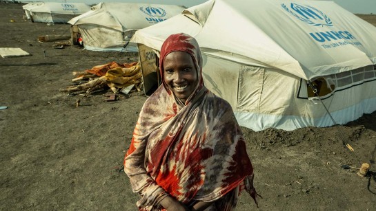 En kvinna på flykt i Sydsudan står framför sitt tält och ler.