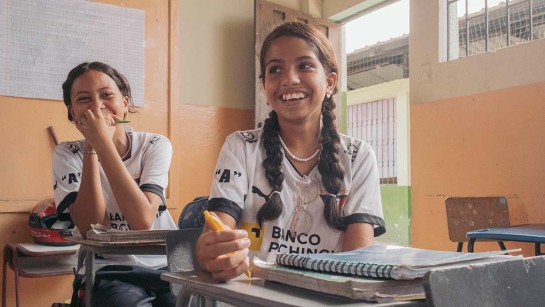 Två glada skolflickor sitter i vid sina bänkar i ett klassrum.