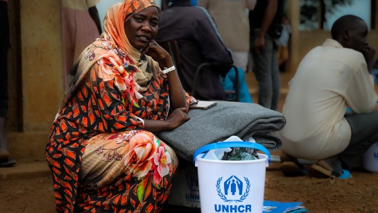 En kvinna sitter på marken och tittar in i kameran. Hon har fått nödhjälp av UNHCR, framför henne ligger filtar och en hink att hämta vatten i. Hon befinner sig på ett mottagningscenter Sydsudan, dit hon precis har anlänt efter att ha flytt från kriget i Sudan.